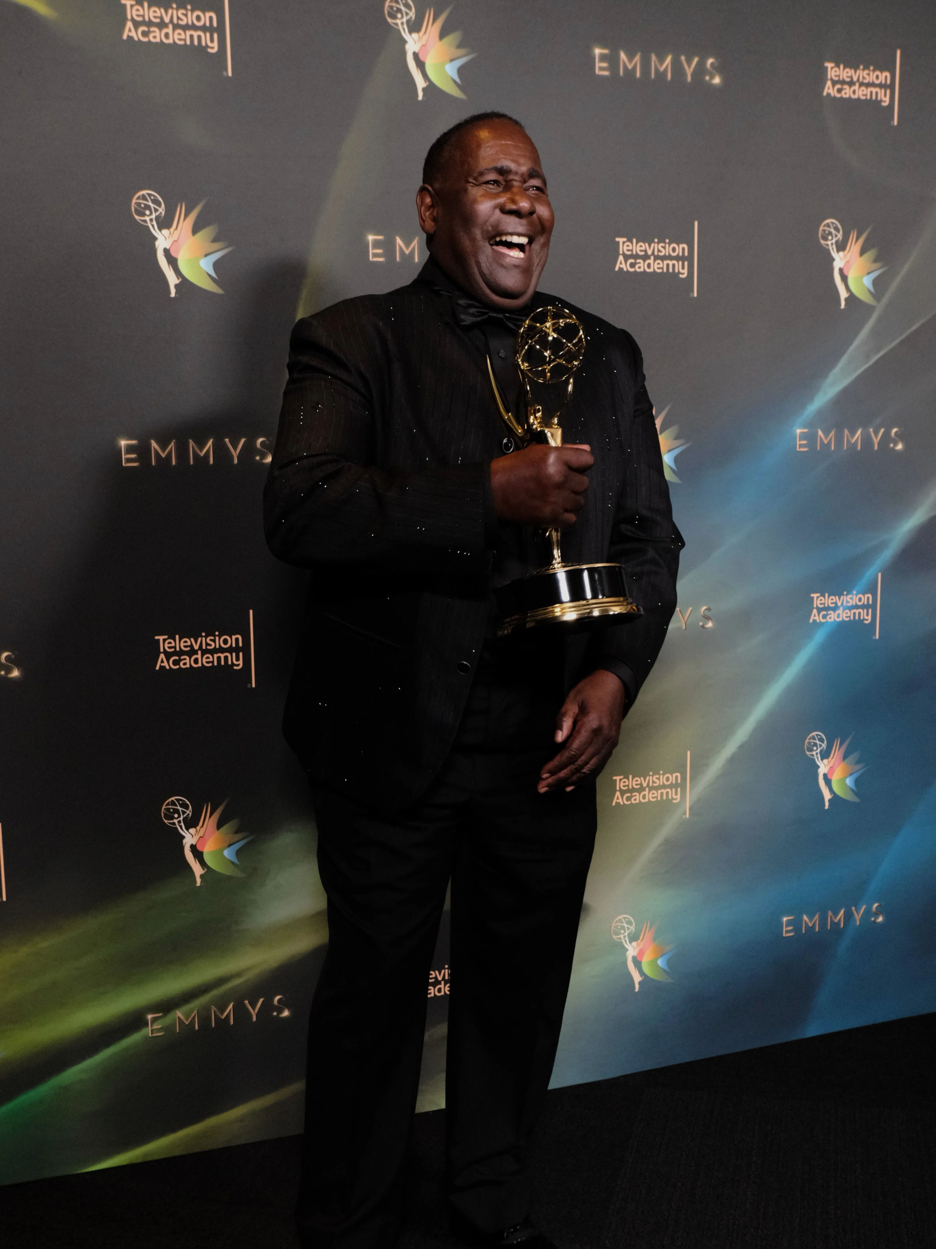 Honoree in peak-lapel black tuxedo holding Emmy with quiet pride