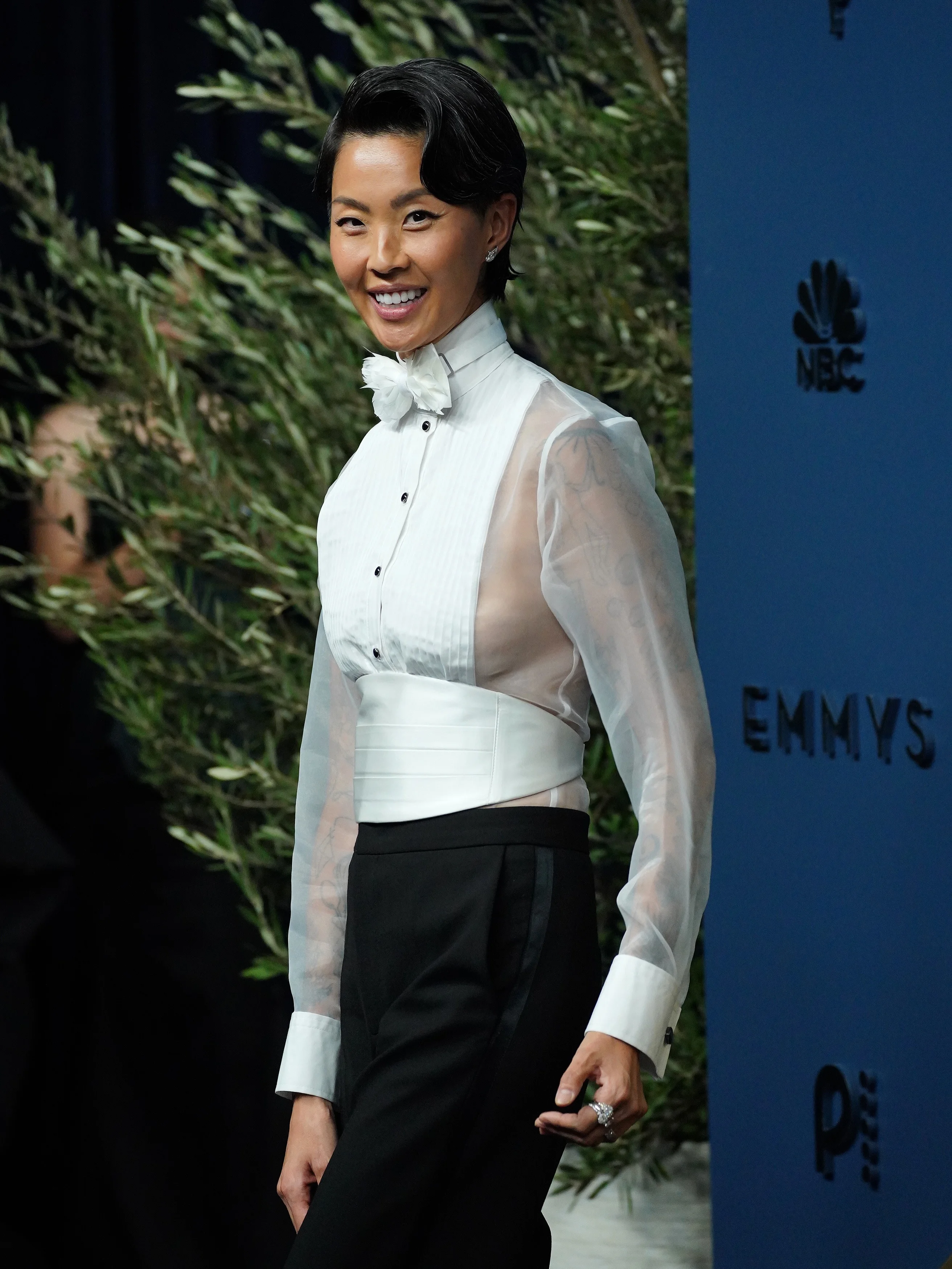 Anna Sawai in sheer white pleated tuxedo blouse and black high-waisted trousers