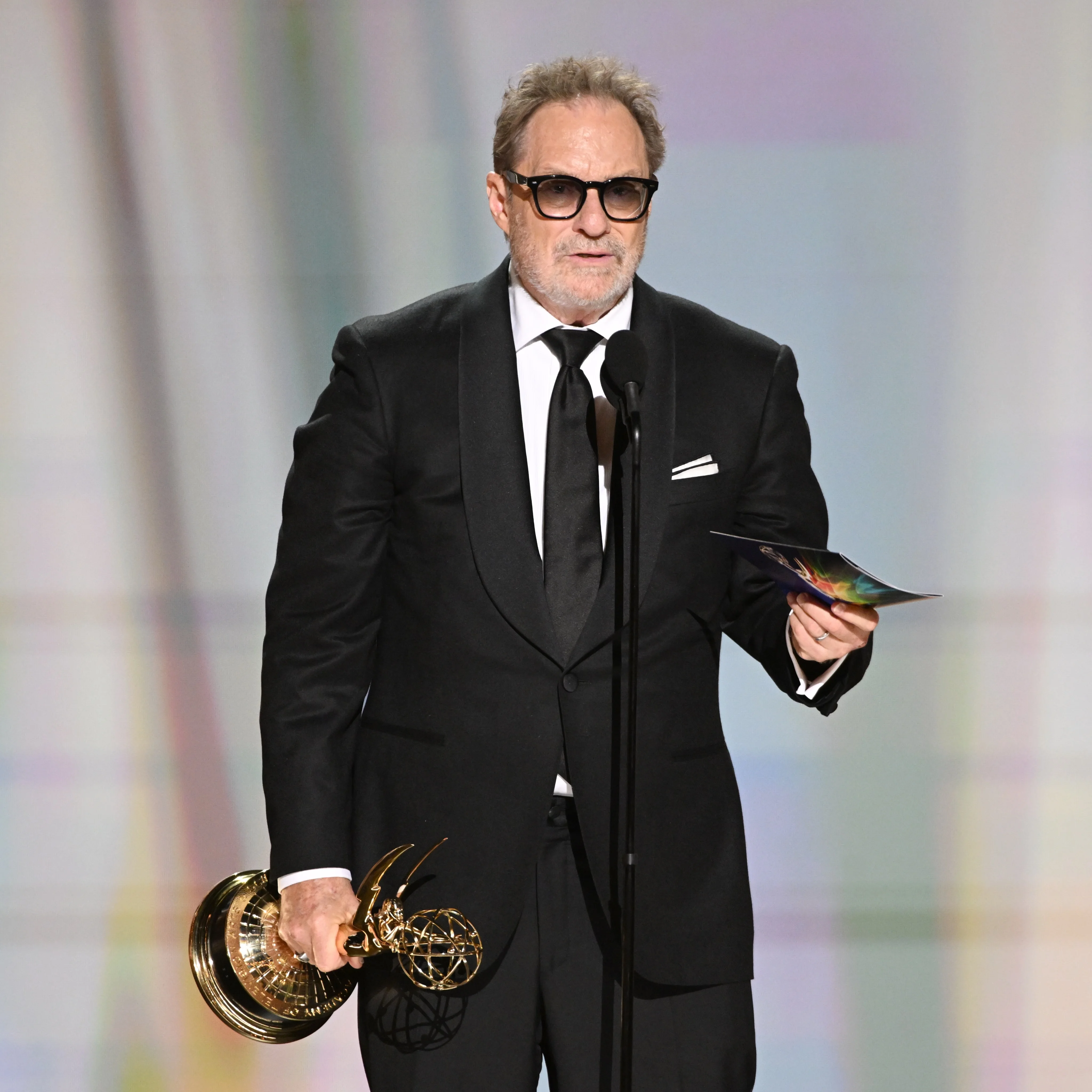 Distinguished creator holding Emmy statuette wearing bold black-rimmed spectacles