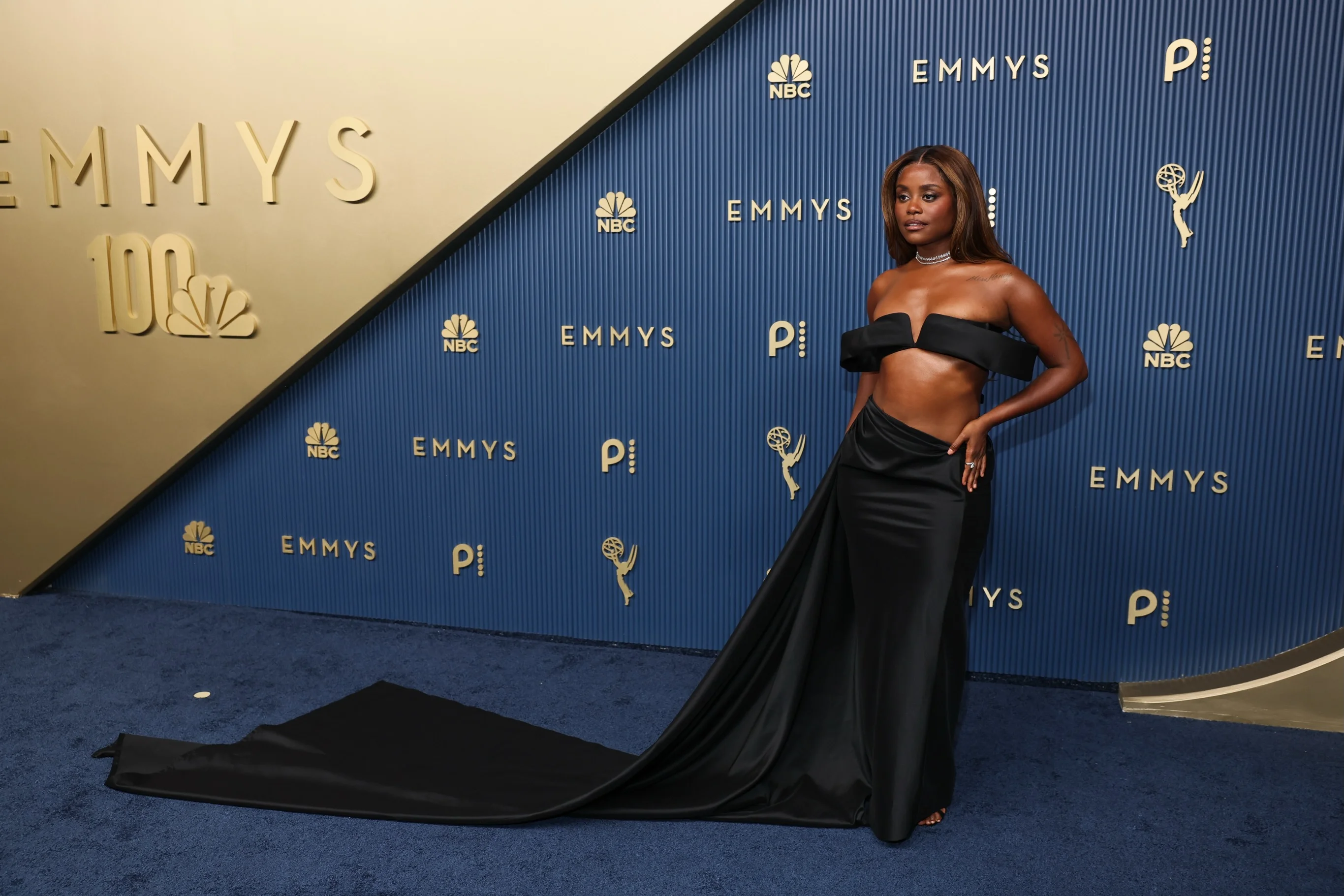 Star posing against gold Emmy logo wall in black gown with cascading train